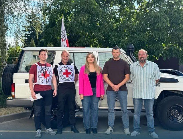 Пятеро людей стоять біля білого автомобіля Червоного Хреста під деревами